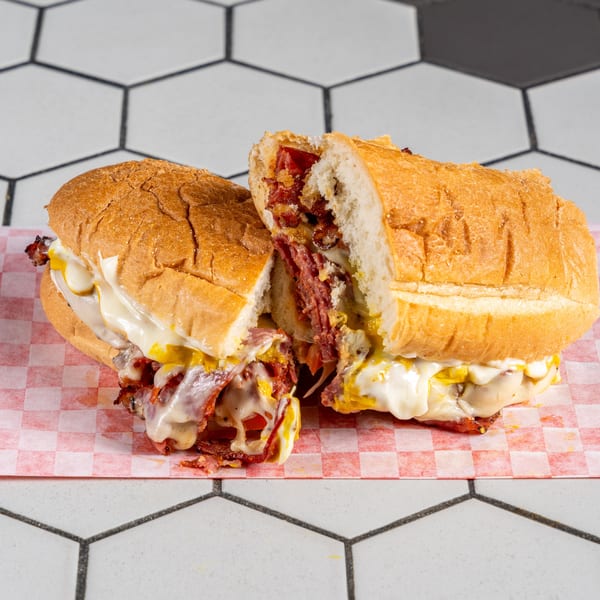 Hearty sub sandwich, sliced in half, with pastrami, melted cheese, and mustard on checkered paper.