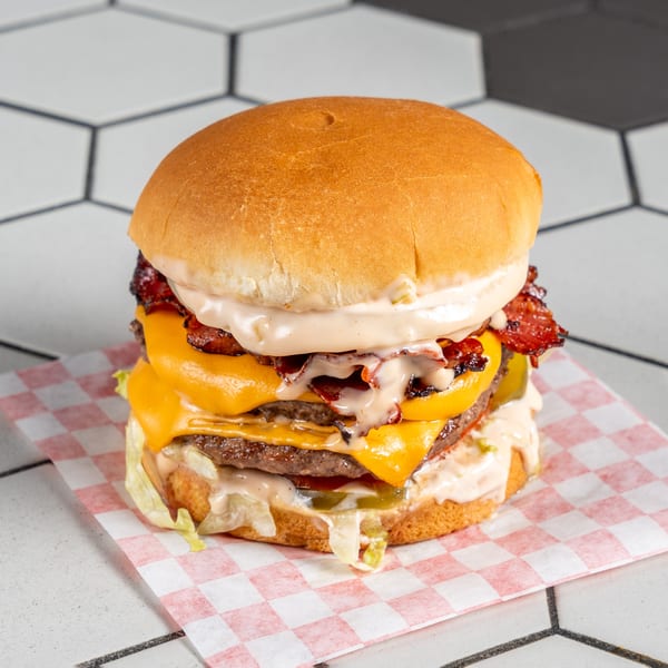 Double cheeseburger with bacon, melted cheese, sauce, lettuce, and pickles on checkered paper.