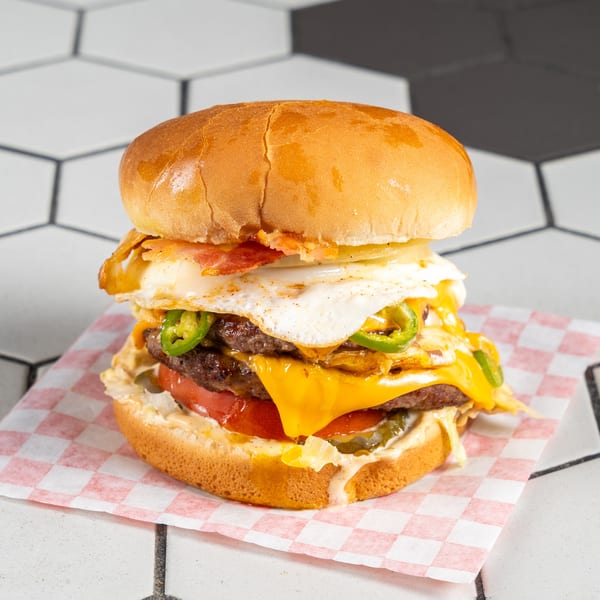 Double patty burger with cheese, fried egg, bacon, jalapeños, and pickles on checkered paper.
