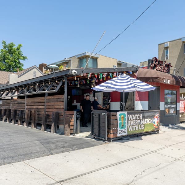 Exterior of The Bucket bar with a wooden patio, international flags, and a striped umbrella.