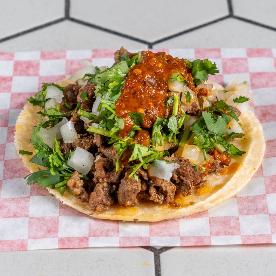 Taco with seasoned meat, diced onions, fresh cilantro, and red salsa on red checkered paper.