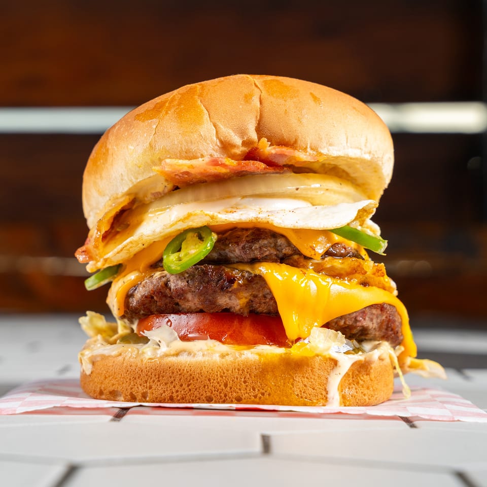 Double cheeseburger stacked with bacon, fried egg, jalapeños, tomato, lettuce, and melted cheese on a bun.