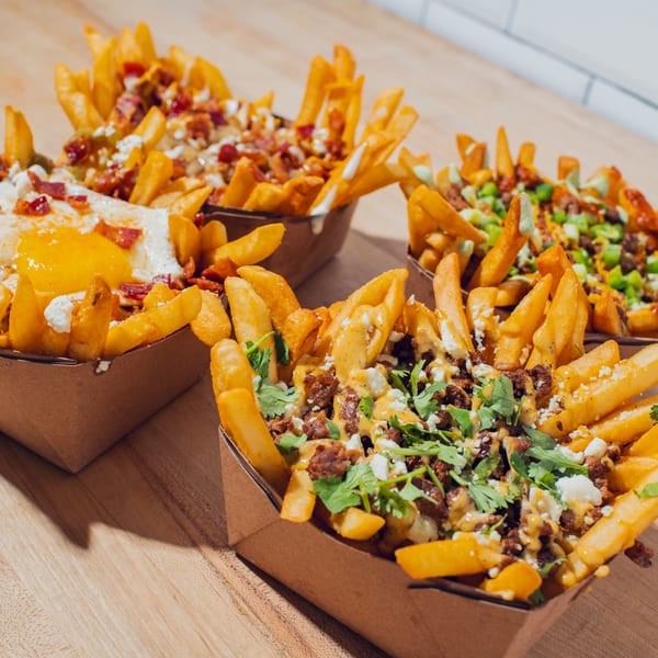 Three trays of loaded fries topped with sauces, cheese, bacon, herbs, vegetables, and a fried egg on a wooden table.