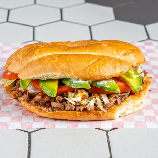 Hearty sub sandwich filled with steak, lettuce, tomato, avocado, and crema on checkered paper.