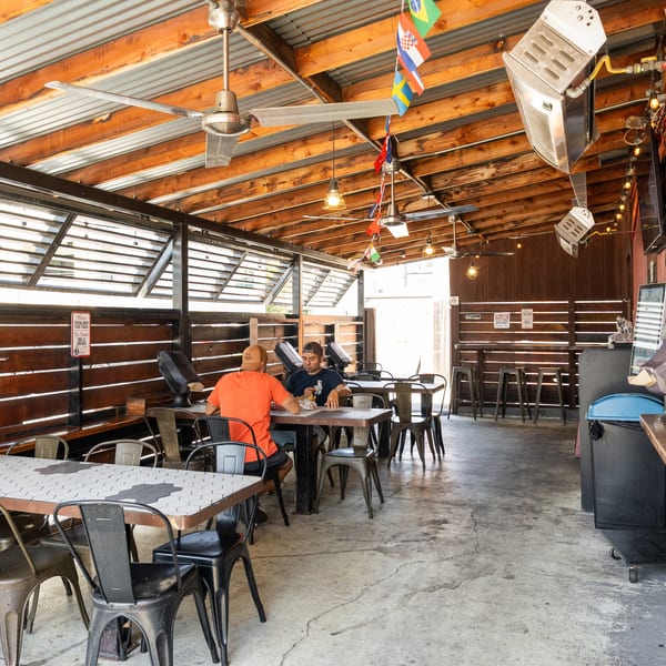 Casual outdoor restaurant patio with two men eating, industrial tables, wooden walls, and hanging flags.
